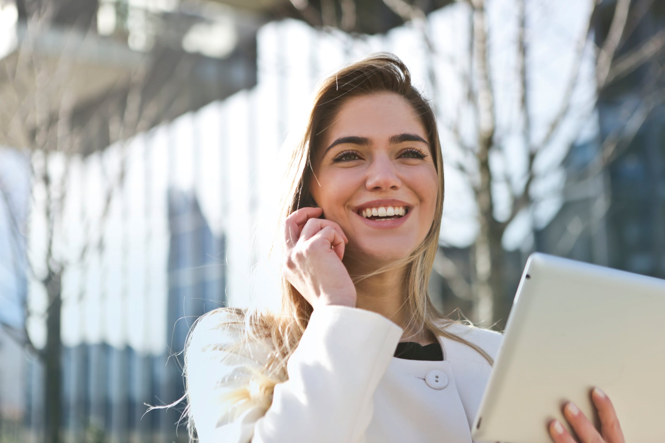 a white woman smiling outside on the phone wearing a white business suit holding a tablet