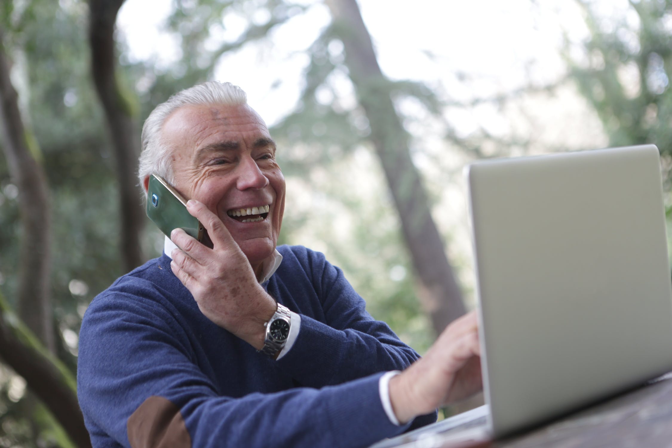 a white senior man wearing a blue sweater on the phone smiling looking at a silver laptop