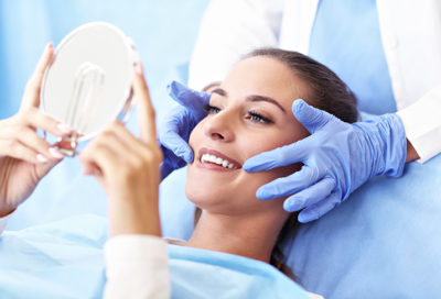 A woman smiles, holding up a mirror while a dentist points out her teeth.