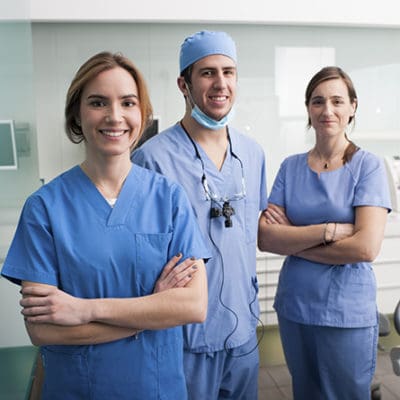 Three dentists, two woman and one male, stand in a dental examination room. They are all wearing blue scrubs