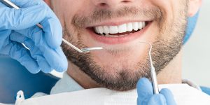 A man with white teeth and blue gloves is holding two tooth brushes.